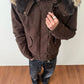 Dark brown washed hooded jacket with plush lined hood and fur trim, hand on the zip, worn with jeans on a wooden floor
