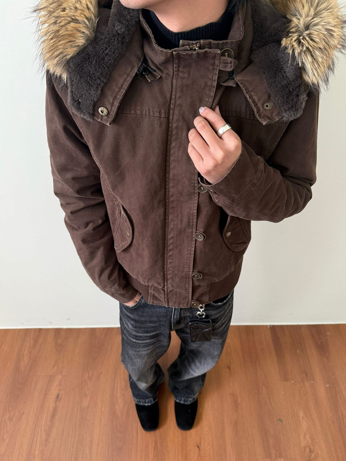 Dark brown washed hooded jacket with plush lined hood and fur trim, hand on the zip, worn with jeans on a wooden floor