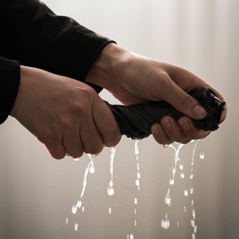 Hands wringing a dark tea cloth as water drips from it against a pale background
