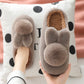 Brown plush bunny slippers with ears and a round pom pom held in a hand over a polka dot cushion, brown variant