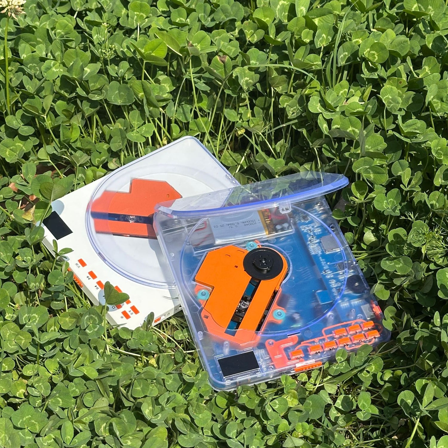 White and clear portable CD players lying open on clover in daylight