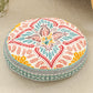 Round cushion with a coral and teal leaf print on a cream top and a teal woven border, on a beige rug