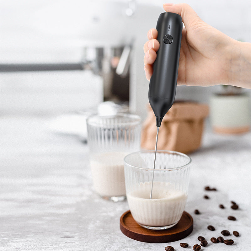 Hand holding a black electric milk frother whisking milk in a glass beside scattered coffee beans