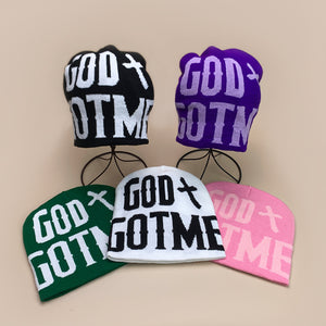 Five knit beanies on stands and a table in black, purple, green, white and pink with large GOD and GOTME lettering