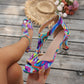 Hand holding a multicolor print T-strap sandal with a straw hat and flowers in the background