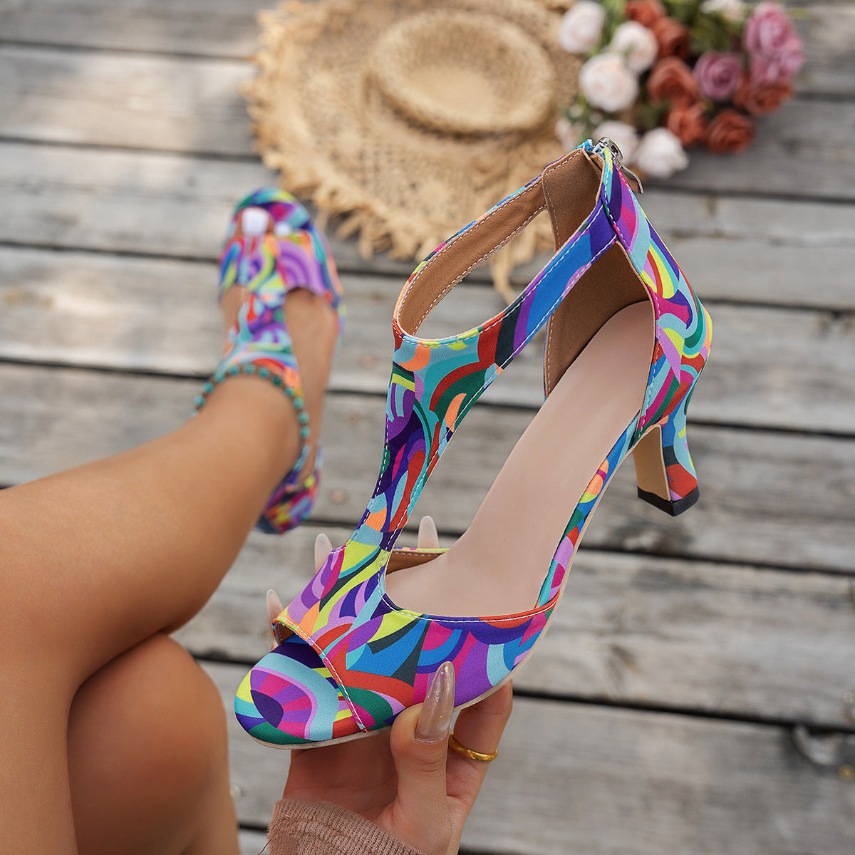 Hand holding a multicolor print T-strap sandal with a straw hat and flowers in the background