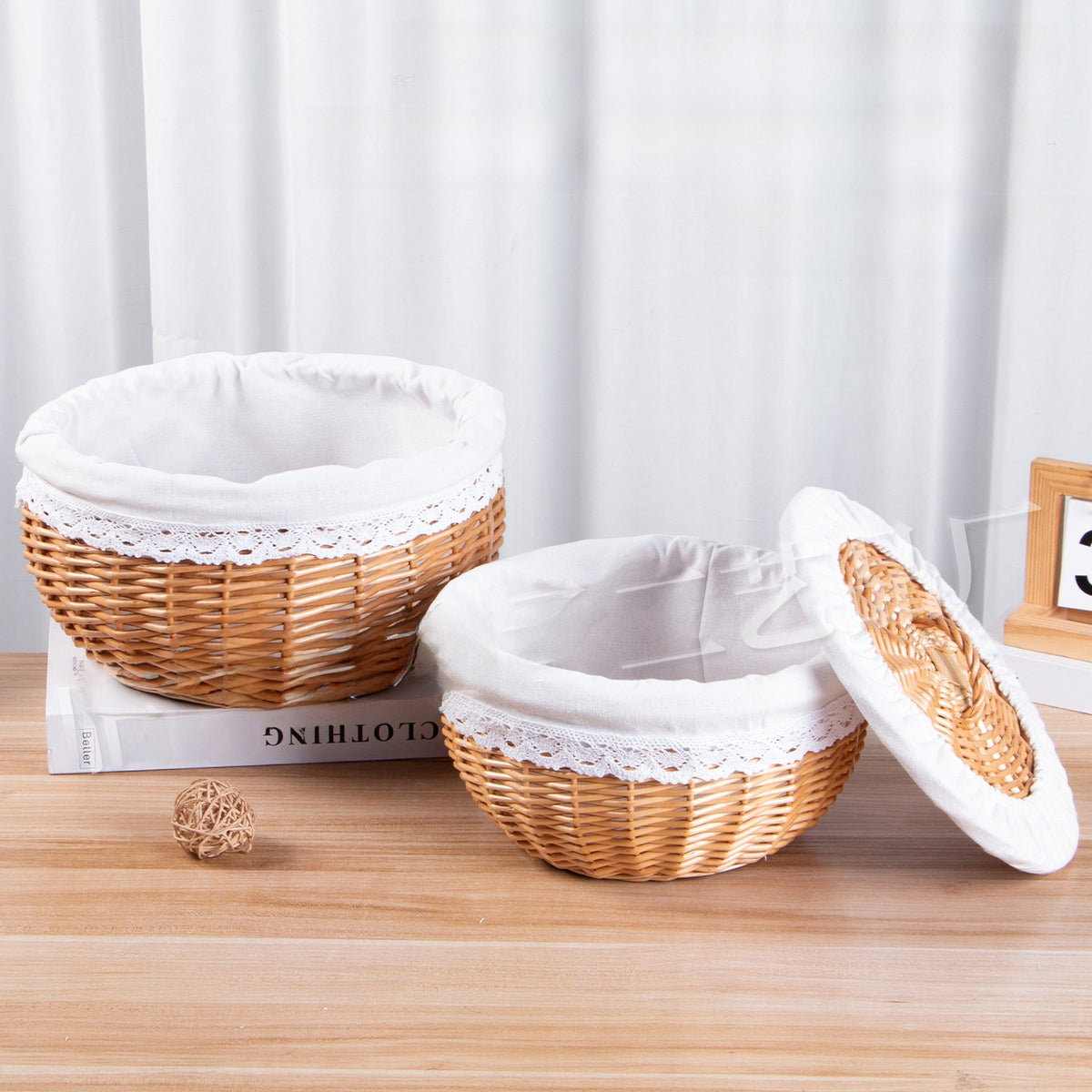 Wicker basket with white lined lace trim open beside its lid on a wooden table with a book and a rattan ball