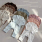 Group of zip front baby rompers in brown, taupe, cream, olive, blue, pink and white laid on a gray floor in sunlight