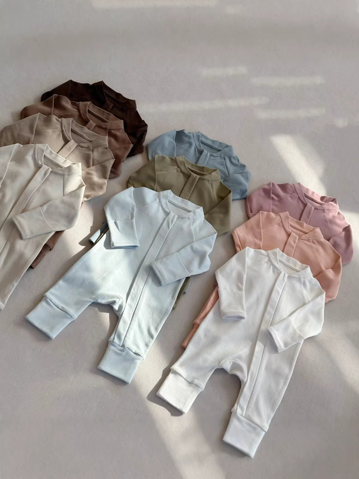 Group of zip front baby rompers in brown, taupe, cream, olive, blue, pink and white laid on a gray floor in sunlight