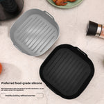 Gray and black silicone air fryer liner trays with ridged bases beside a plate of cooked bacon