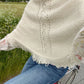 Cream knit poncho top with fringe hem worn over a floral blouse and jeans on a bicycle beside a green field