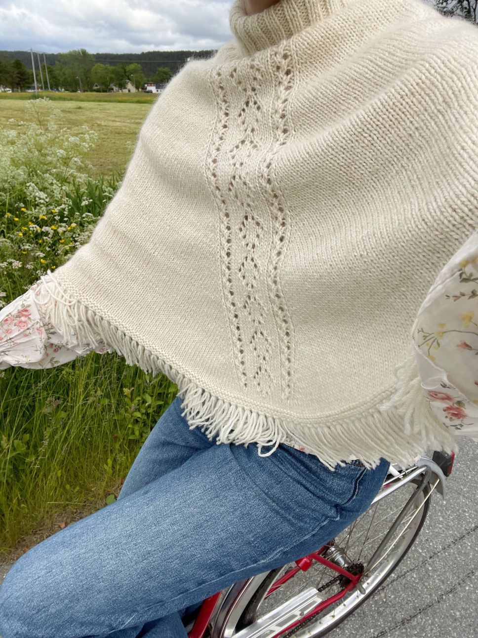 Cream knit poncho top with fringe hem worn over a floral blouse and jeans on a bicycle beside a green field