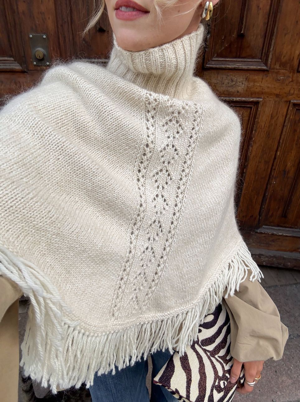 Cream knit poncho top with a high ribbed collar, openwork panel and fringe hem in front of a wooden door
