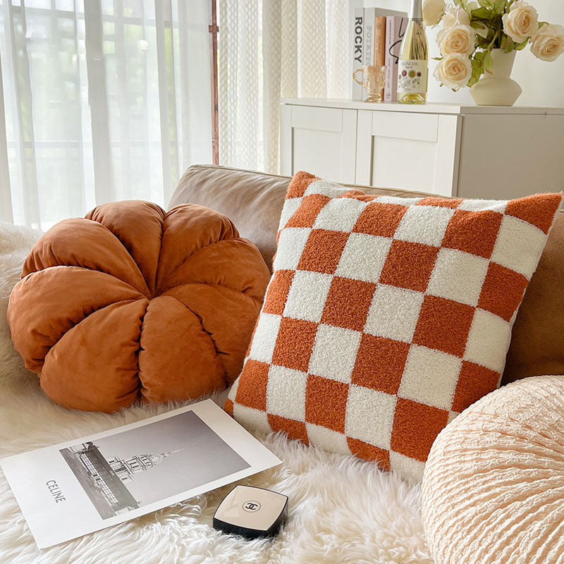Orange pumpkin pillow and orange and cream checkerboard plush pillow on a sofa with a white throw in a bright room