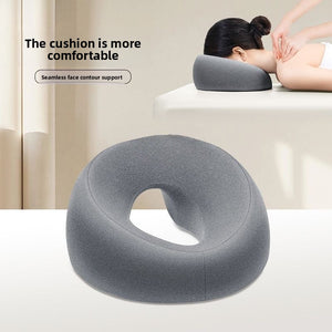 Gray U-shaped memory foam face rest pillow with an open center, shown on a table beside a person lying face down resting on it