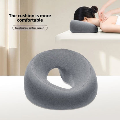 Gray U-shaped memory foam face rest pillow with an open center, shown on a table beside a person lying face down resting on it