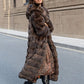 Woman in a long brown hooded fur coat turning mid step on a city street, printed dress and boots showing