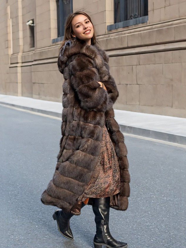 Woman in a long brown hooded fur coat turning mid step on a city street, printed dress and boots showing