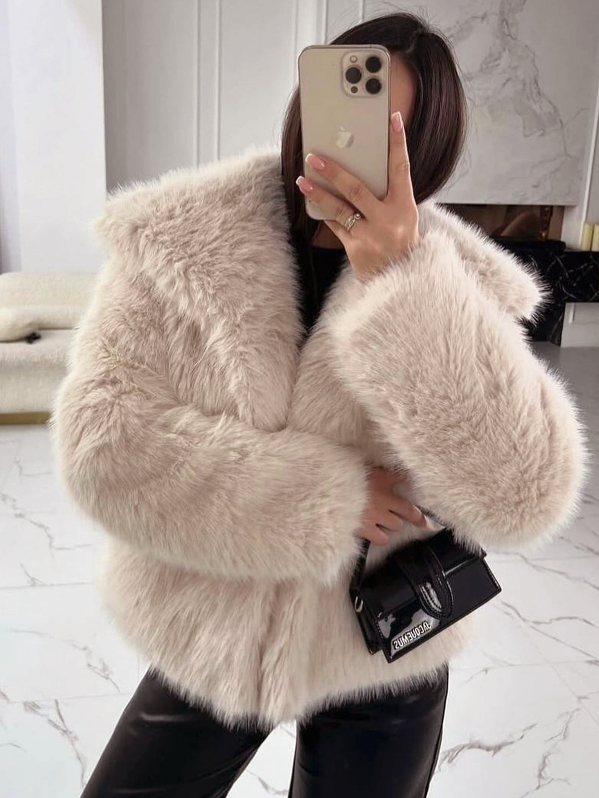 Woman in a pale beige vegan fur jacket taking a mirror selfie in black trousers with a small black bag