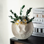 White glazed ceramic cowboy hat vase holding daisies and eucalyptus on a dark shelf