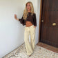 Model leaning on a white wall beside a wooden door, wearing beige wide leg trousers with a cuffed hem