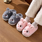 Two pairs of cartoon bunny plush slippers in slate gray and pink on a wooden floor next to a foot in a white fleece trouser