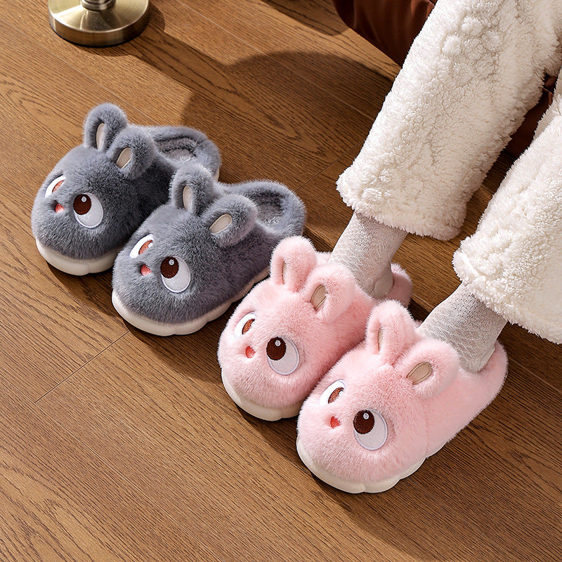 Two pairs of cartoon bunny plush slippers in slate gray and pink on a wooden floor next to a foot in a white fleece trouser
