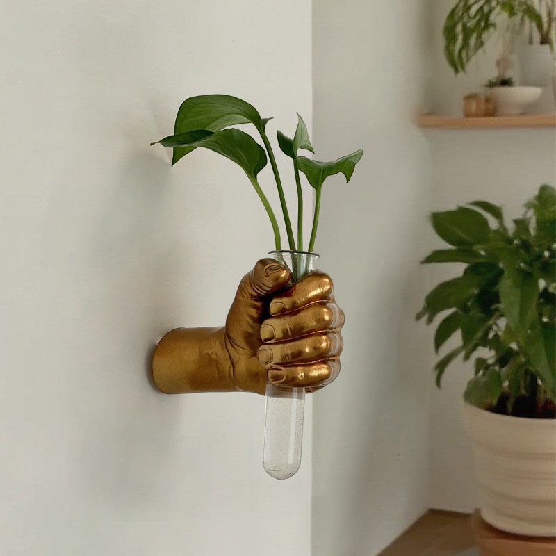 Gold hand shaped wall vase gripping a glass test tube with a green stem on a white wall