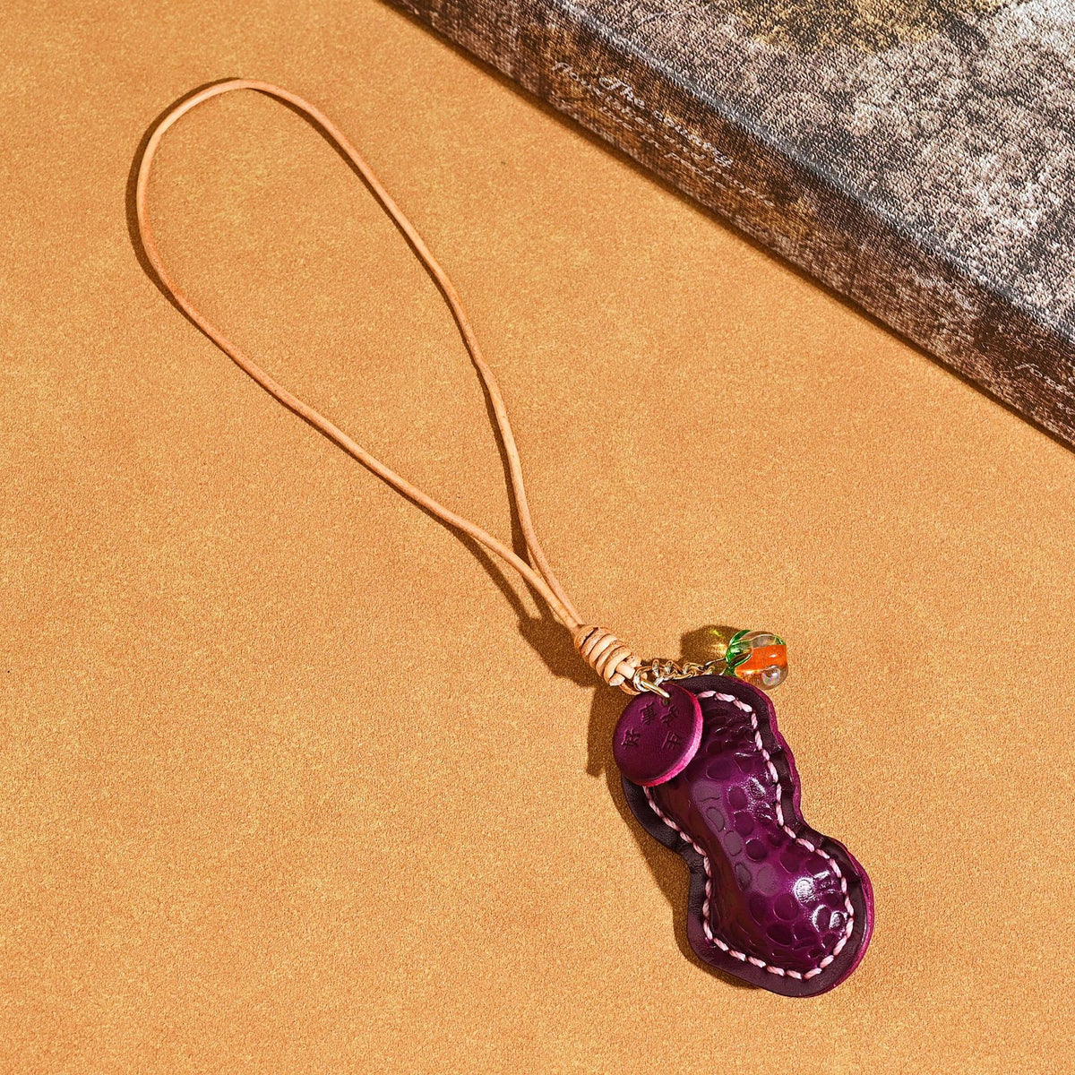 Purple peanut shaped leather pendant with a leather cord loop, orange glass bead and stamped tag on a tan background