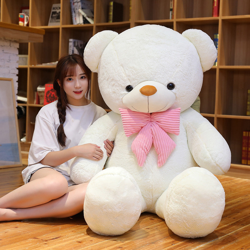 Cream white plush teddy bear with a pink striped bow tie and a woman sitting beside it in front of a bookshelf