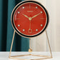 Orange dial desk clock with black bezel and gold hands on a triangular gold stand with a swinging gold ball, teal curtain