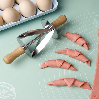 Stainless steel croissant roller cutter with wooden handle on a dough mat beside cut croissant dough pieces