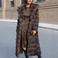 Woman smiling in a long brown hooded fur coat with banded pile, standing on a street beside a stone wall