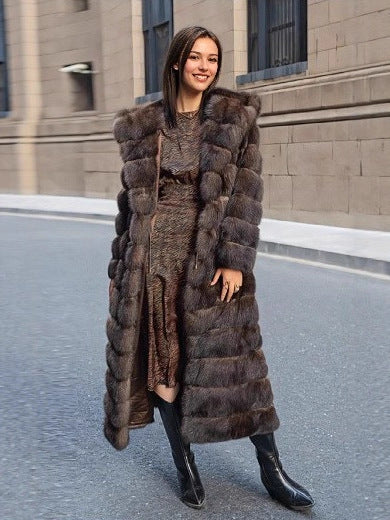 Woman smiling in a long brown hooded fur coat with banded pile, standing on a street beside a stone wall