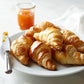 Plate of golden baked croissants with a jar of jam and a knife on a white table