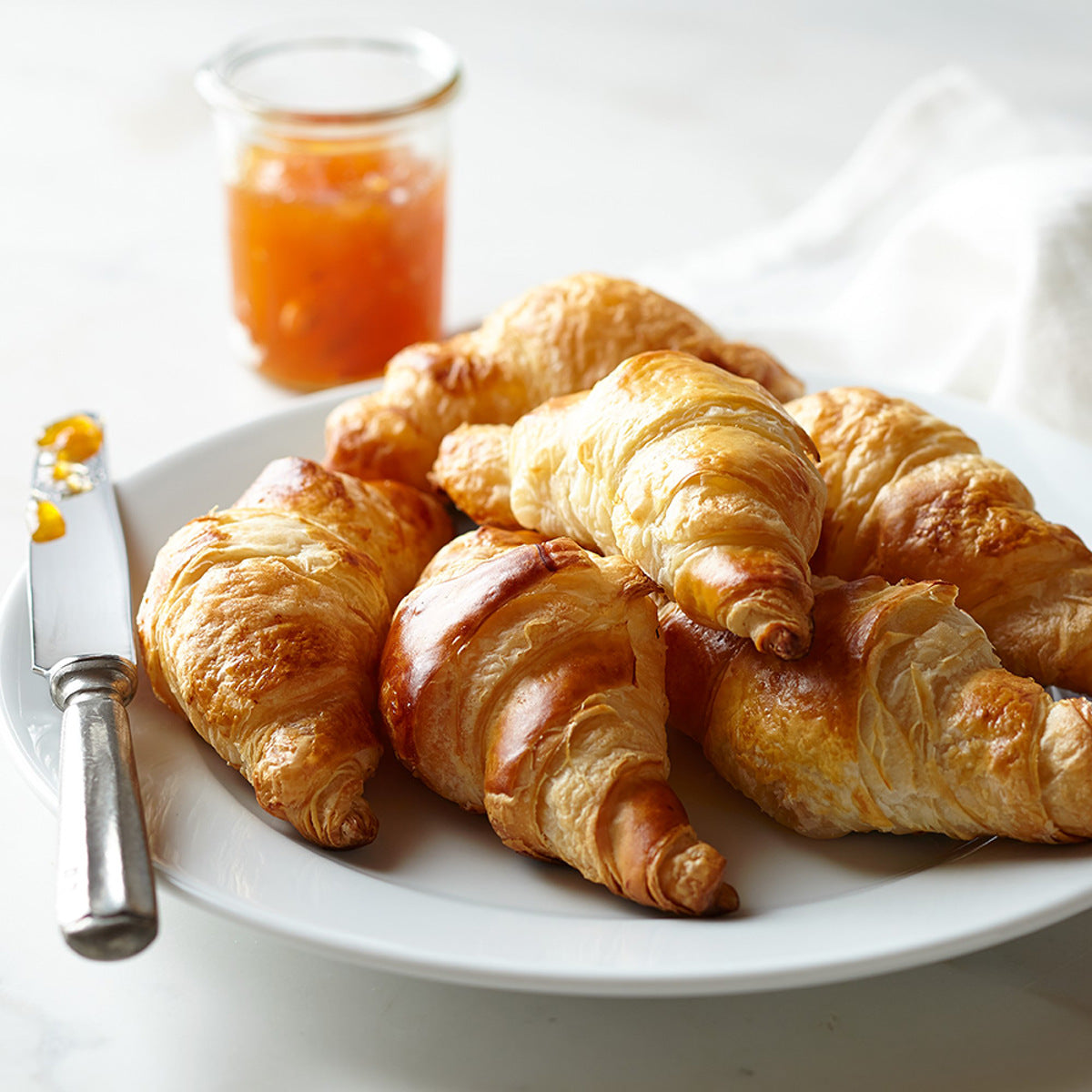 Plate of golden baked croissants with a jar of jam and a knife on a white table