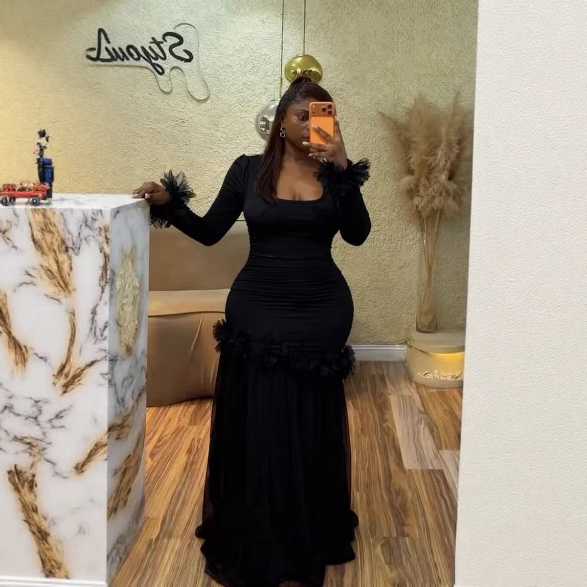 Woman in a black ruched maxi dress with rosette trim at the cuffs and the hip, hand resting on a marble pillar