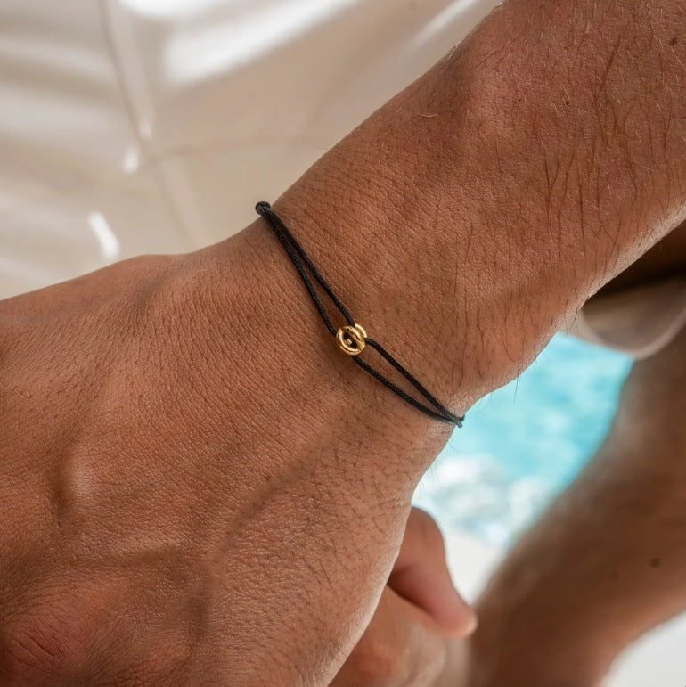 Thin black cord lucky charm bracelet with a small gold charm worn on a mans wrist