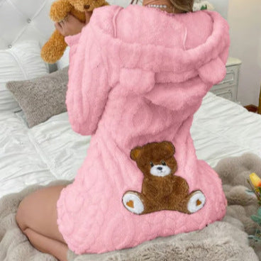 Back view of the pink plush hooded romper with bear ears and a teddy bear patch, worn kneeling on a bed