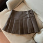 Coffee brown pleated mini skirt with two buckle straps on the side of the yoke, laid flat on a sofa