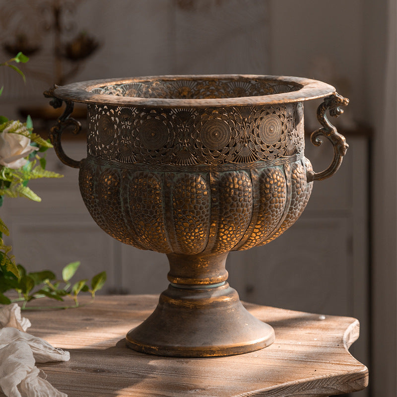 Vintage iron pedestal urn with two scroll handles and aged bronze gold finish on a wooden table