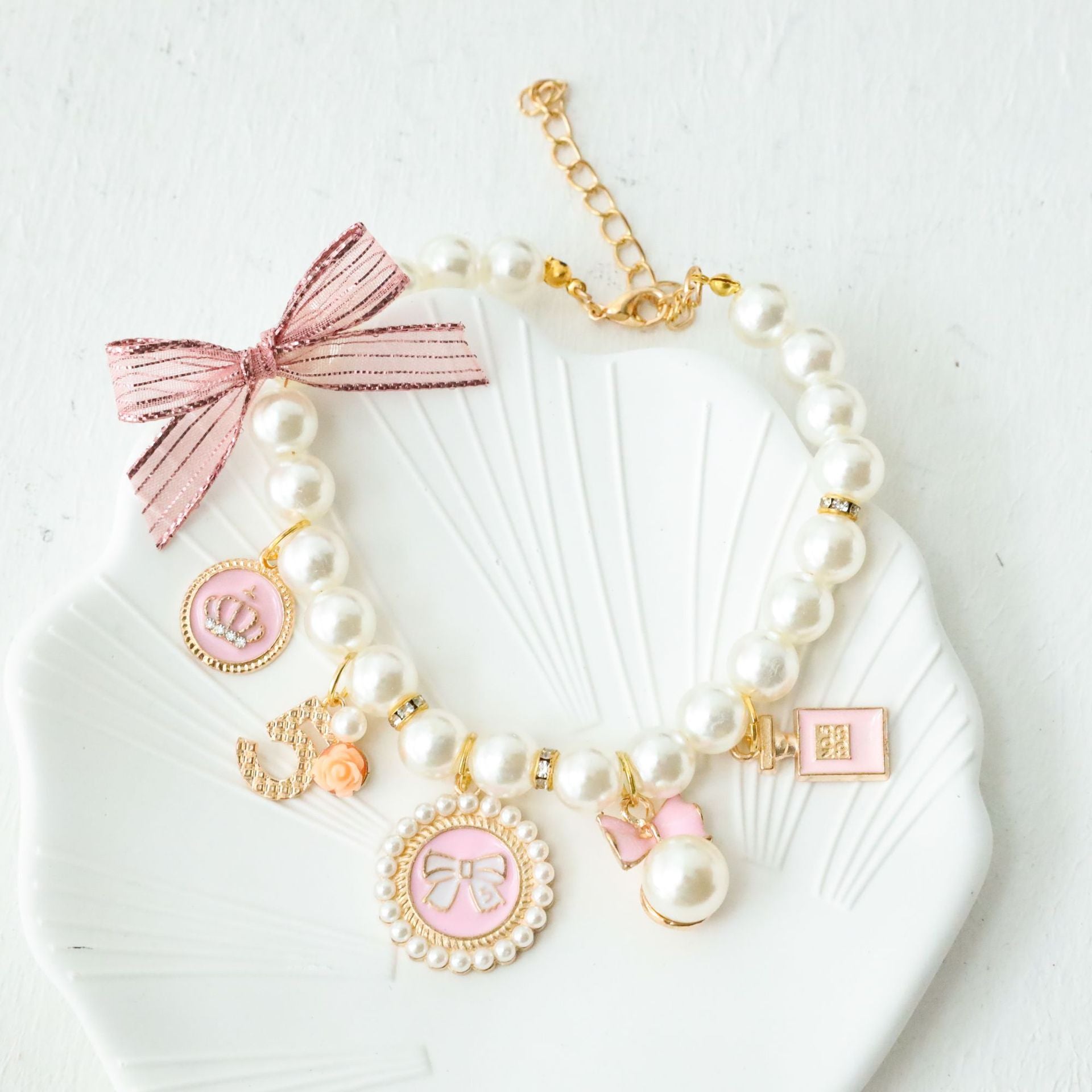 Powder pink faux pearl cat collar with charm pendants and an organza ribbon bow on a shell dish