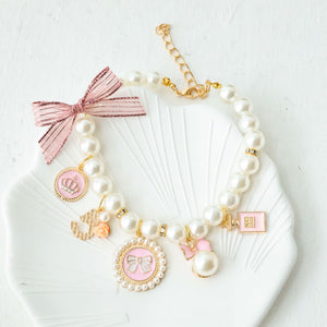 Powder pink faux pearl cat collar with charm pendants and an organza ribbon bow on a shell dish