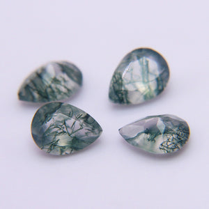 Four pear cut moss agate stones on a pale grey surface, green dendrites under faceted crowns