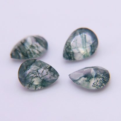 Four pear cut moss agate stones on a pale grey surface, green dendrites under faceted crowns