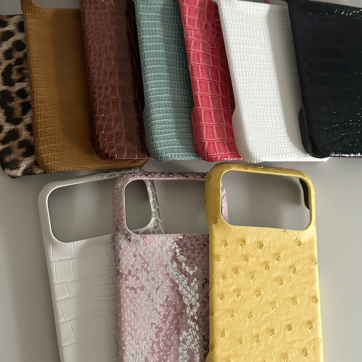 Fanned stack of leopard, tan, brown, teal, coral, white and black phone cases above white, pink snake and yellow cases