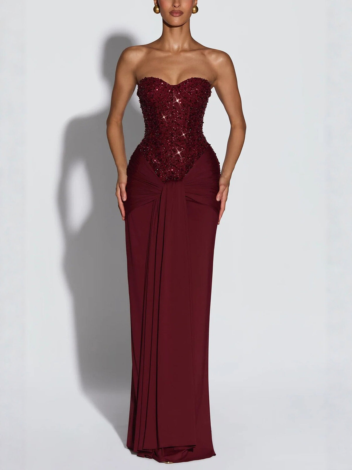 Woman in a burgundy strapless long dress with a sequin bodice and draped hip knot on a grey background
