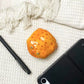 Plain orange resin frog earbud case seen from above on white cloth beside a pen and a tablet