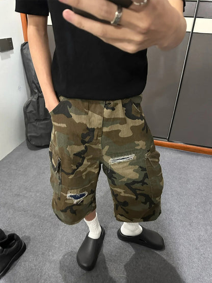 Close view of the camo cargo shorts on a model in a black tee, hand in the pocket, a rip on the front of the leg
