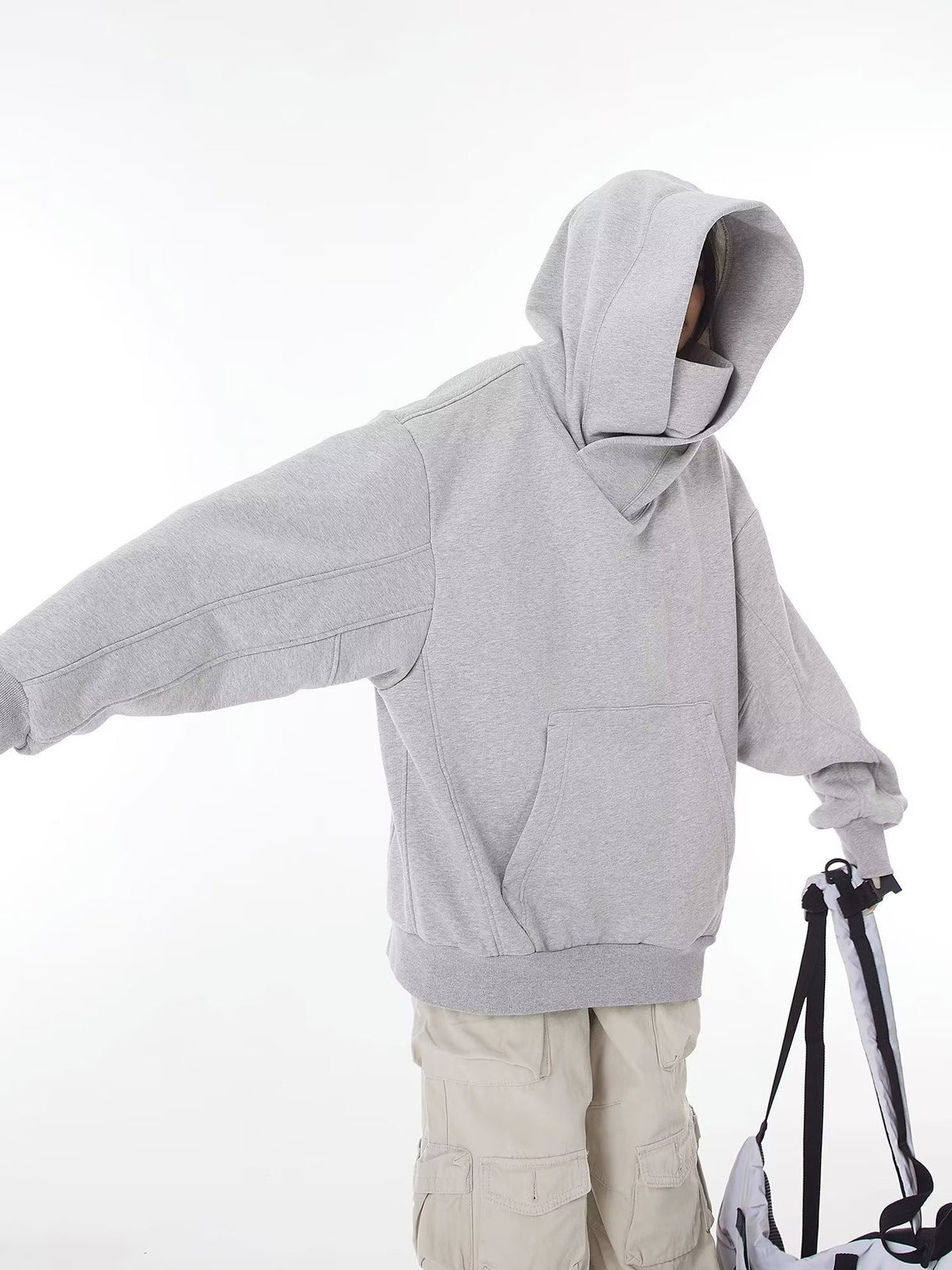 Grey hooded sweatshirt with the hood up and one arm extended, showing the front pocket and ribbed hem, cream cargo trousers